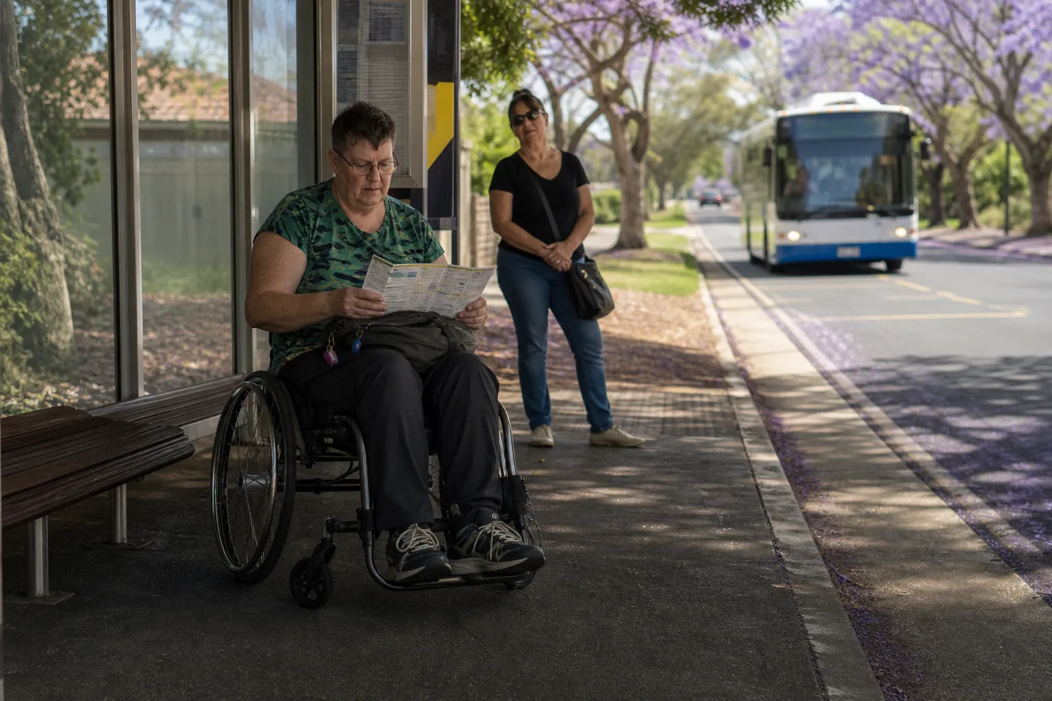 Illustrative wheelchair user reviewing a folded activity plan at a shaded Brisbane bus stop as a bus approaches; a support worker waits nearby.